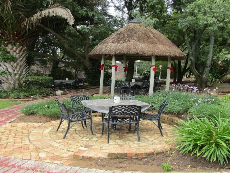 A landscaped garden area featuring a circular brick paved patio, mature trees, and a traditional thatched-roof gazebo for outdoor seating.
