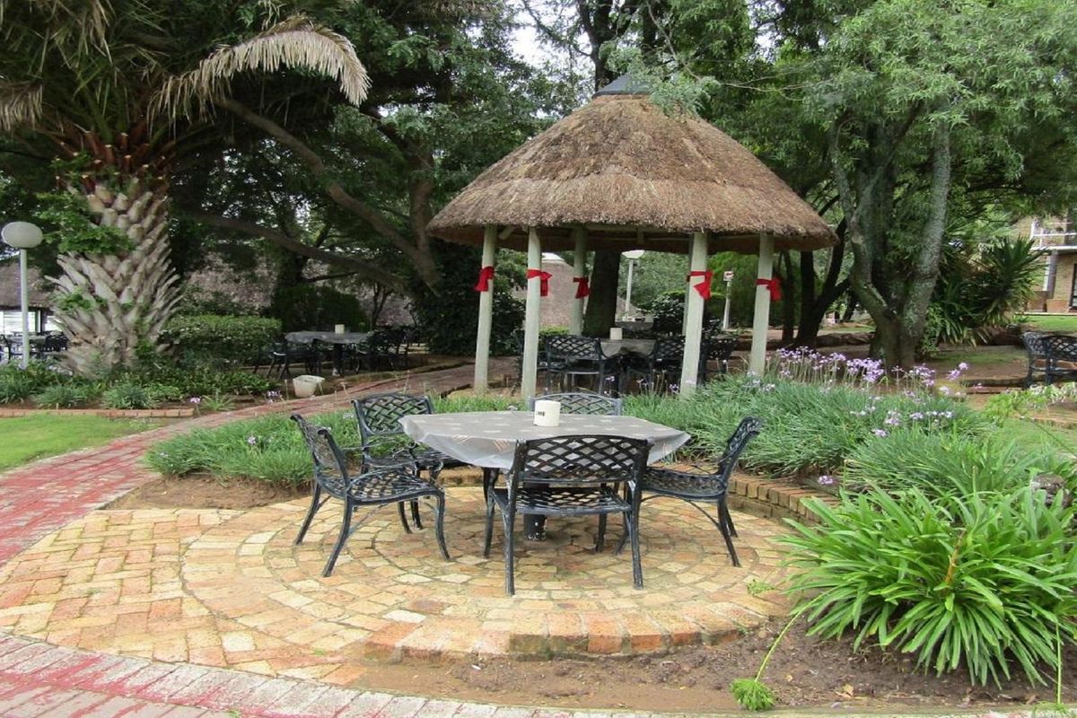 A landscaped garden area featuring a circular brick paved patio, mature trees, and a traditional thatched-roof gazebo for outdoor seating.