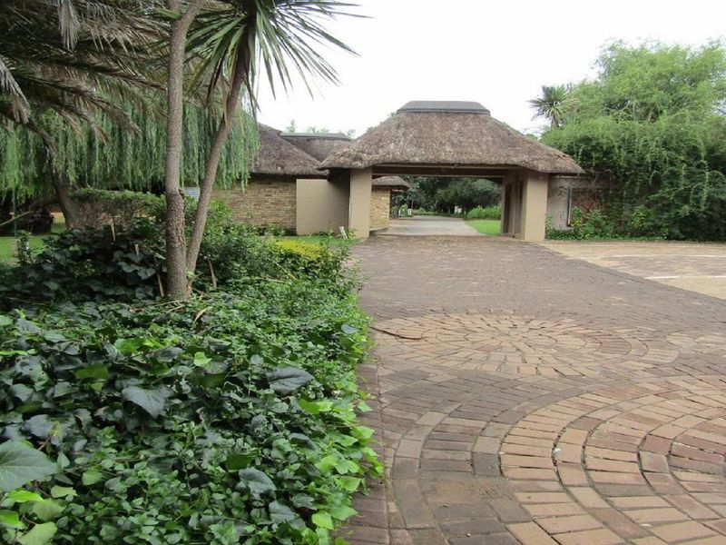 A paved driveway leading to a thatched-roof entrance structure, surrounded by established garden landscaping and mature trees.