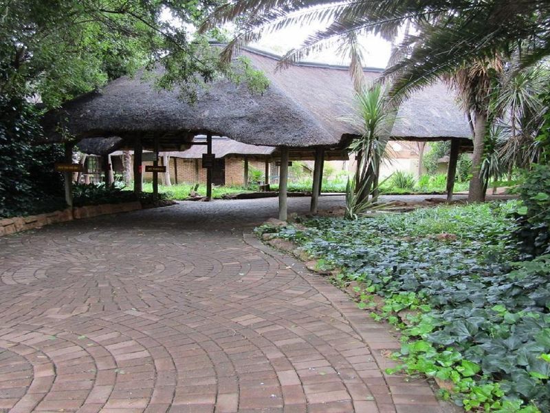 A paved driveway leading to a covered entrance area featuring a traditional thatch roof structure surrounded by mature garden landscaping.