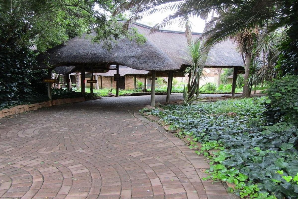 A paved driveway leading to a covered entrance area featuring a traditional thatch roof structure surrounded by mature garden landscaping.