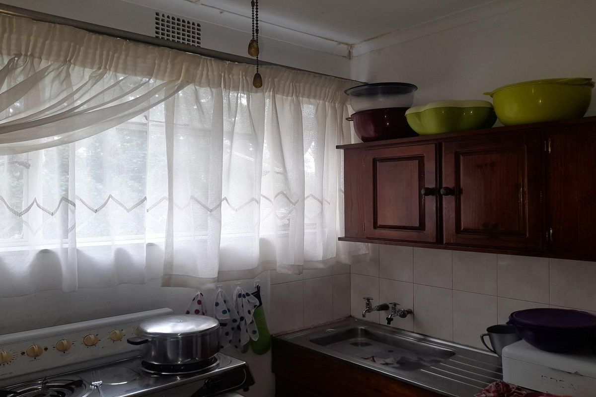 A dated kitchen featuring wooden cabinetry, a stainless steel sink, and a built-in stove, situated beneath a window with sheer curtains.