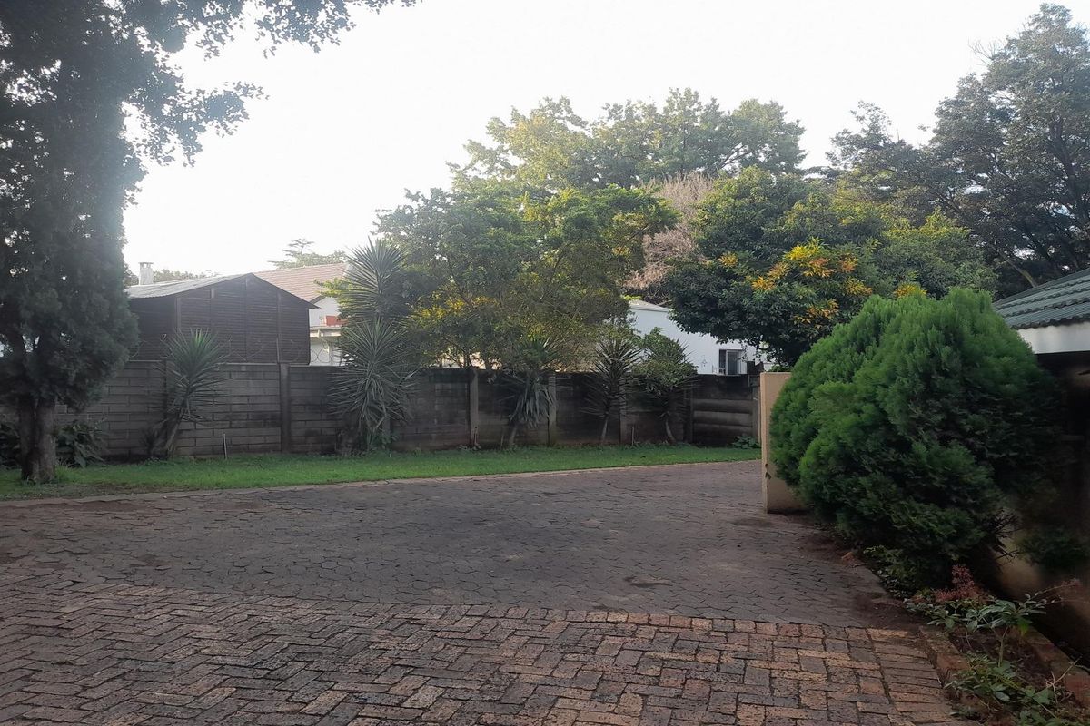 A view of the property's rear garden area featuring a paved driveway, established trees, and a perimeter boundary wall.