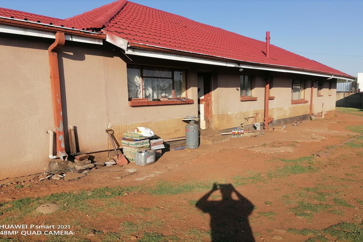 Rear exterior view of a single-story house featuring a red tiled roof, plastered walls, and exposed downpipes, set against an unpaved backyard area.