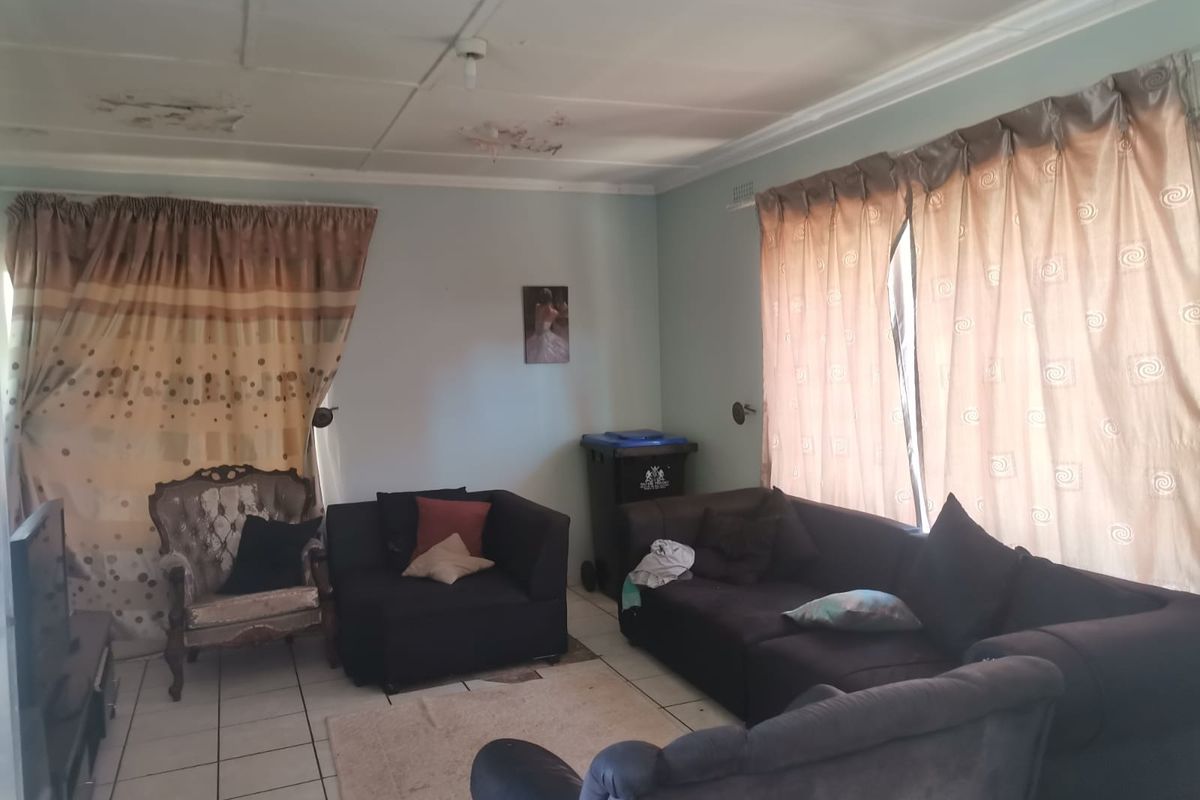 A living room featuring tiled flooring and large windows with curtains. The ceiling shows visible signs of water damage.