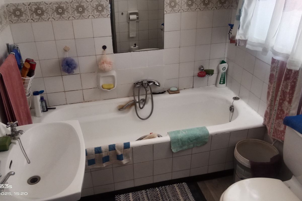A compact bathroom featuring a standard bathtub with handheld shower mixer, pedestal basin, and toilet, finished with dated wall tiling.