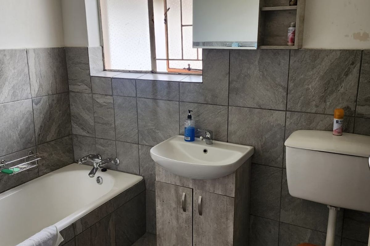 A functional bathroom featuring grey stone-look wall tiles, a built-in bathtub, a vanity basin with storage, and a standard toilet.