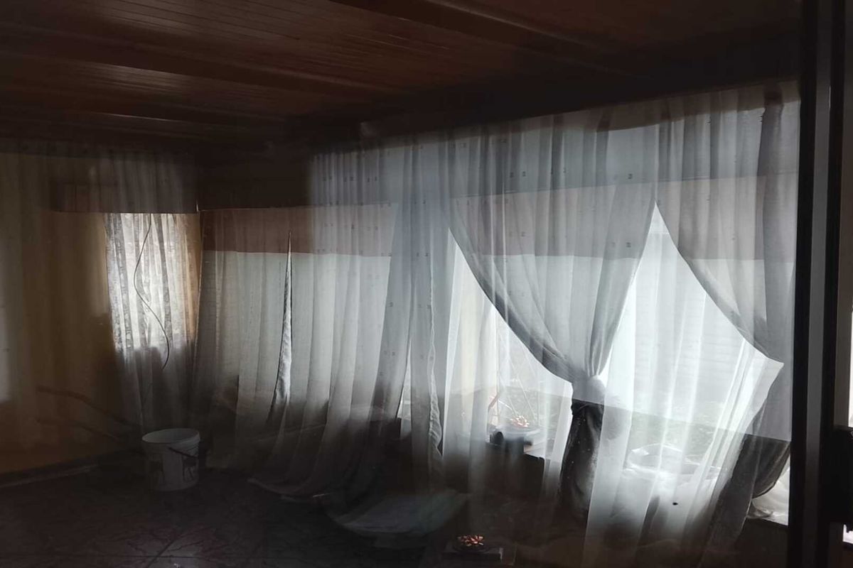 A dimly lit living area featuring a timber-clad ceiling and large windows with sheer curtains, offering a dated aesthetic ready for modernization.