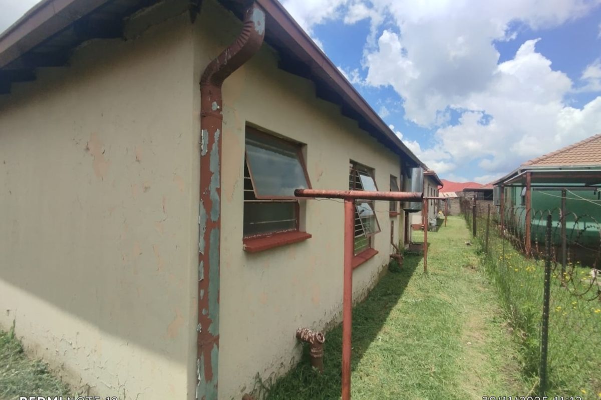 Side exterior view of the house showing weathered wall finishes, rusted downpipes, and metal window frames along a narrow garden path.