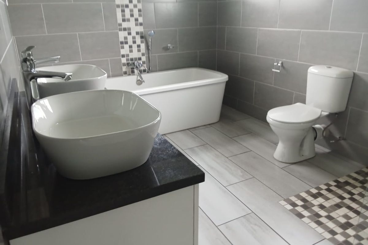 Modern bathroom featuring dual vessel sinks on a dark vanity, a freestanding bathtub, and a separate toilet, finished with contemporary grey wall and floor tiles.