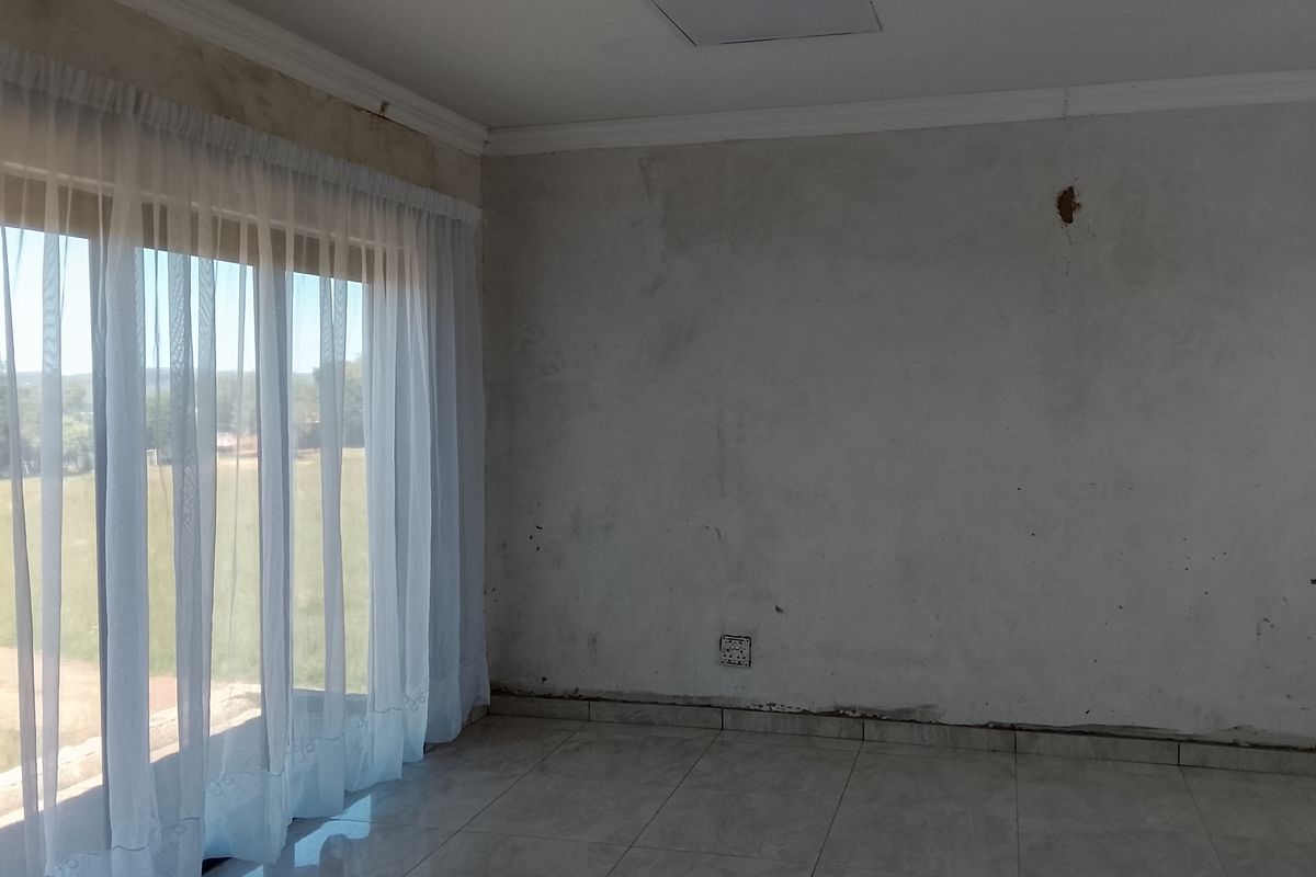 An empty living room featuring tiled flooring, large windows with sheer curtains, and walls requiring plastering and finishing work.