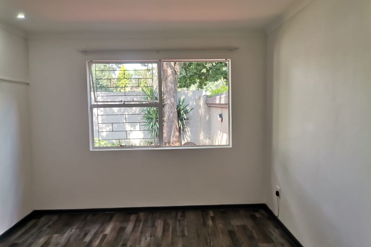 A bright, empty bedroom featuring modern wood-look laminate flooring, neutral walls, and a window with security bars overlooking the garden.