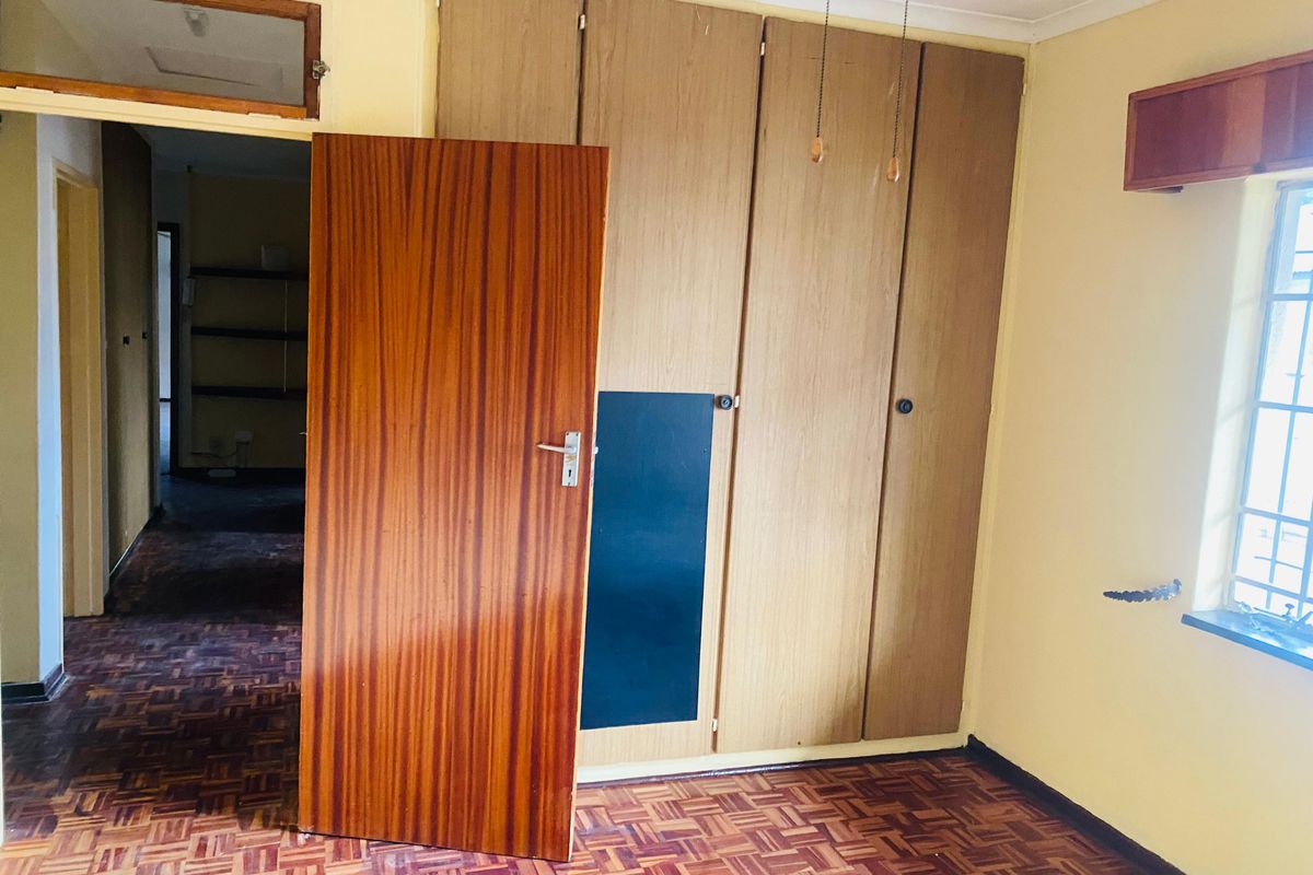 A bedroom featuring original parquet flooring, built-in wooden cupboards, and a window with a metal security frame, leading into a hallway.