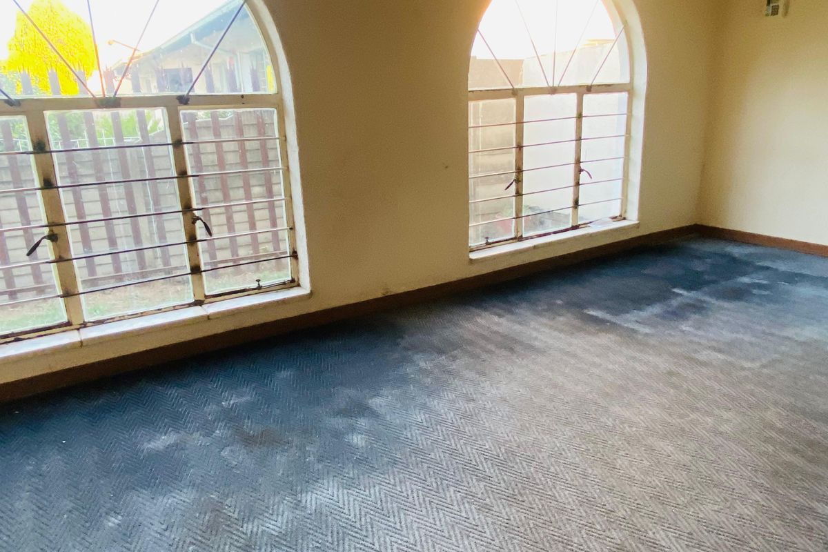 An empty bedroom featuring arched windows with security bars and worn carpet flooring, offering a blank canvas for renovation.