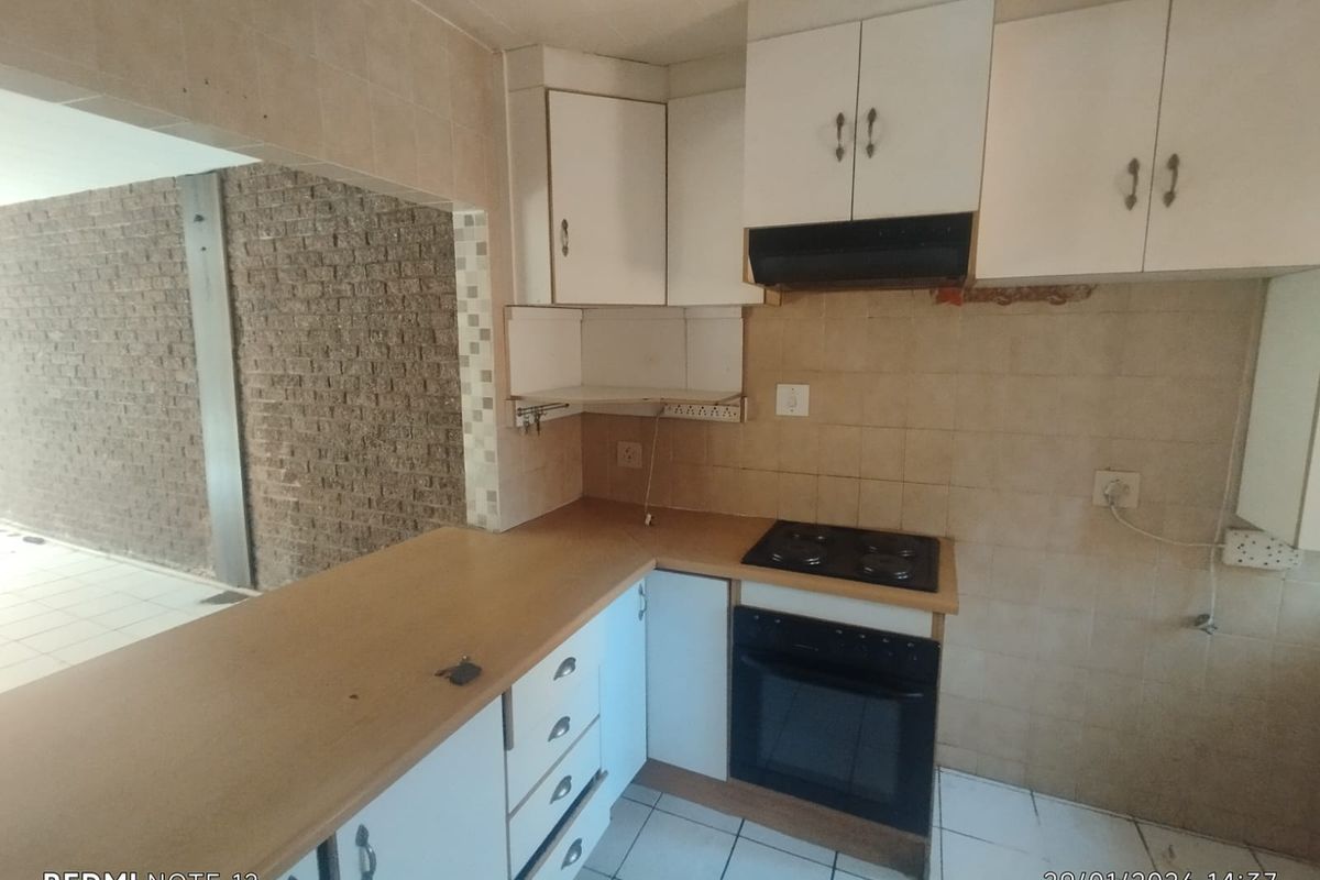 A compact kitchen featuring white cabinetry, a built-in oven and hob, and tiled flooring, opening into an adjacent living area with a brick feature wall.