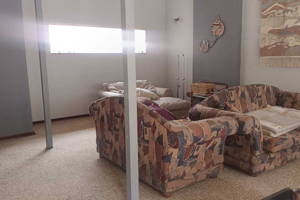A living room area featuring carpeted flooring, high ceilings, and exposed structural support columns, providing a dated interior space with potential for modernization.