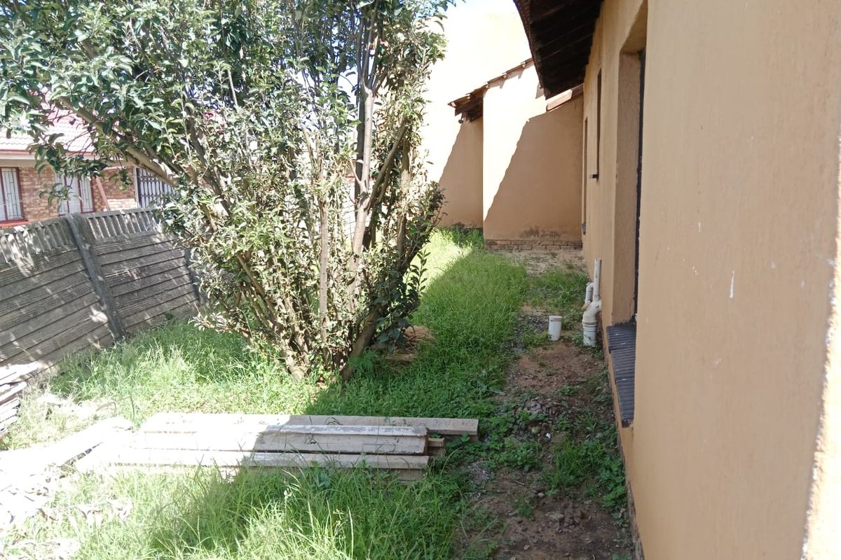 A side exterior view of the property featuring a narrow garden area with overgrown grass and exposed plumbing infrastructure along the wall.