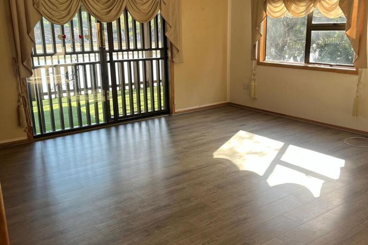 A spacious, sunlit living room featuring modern wood-look laminate flooring, large windows, and a sliding glass door with security gate access to the exterior.