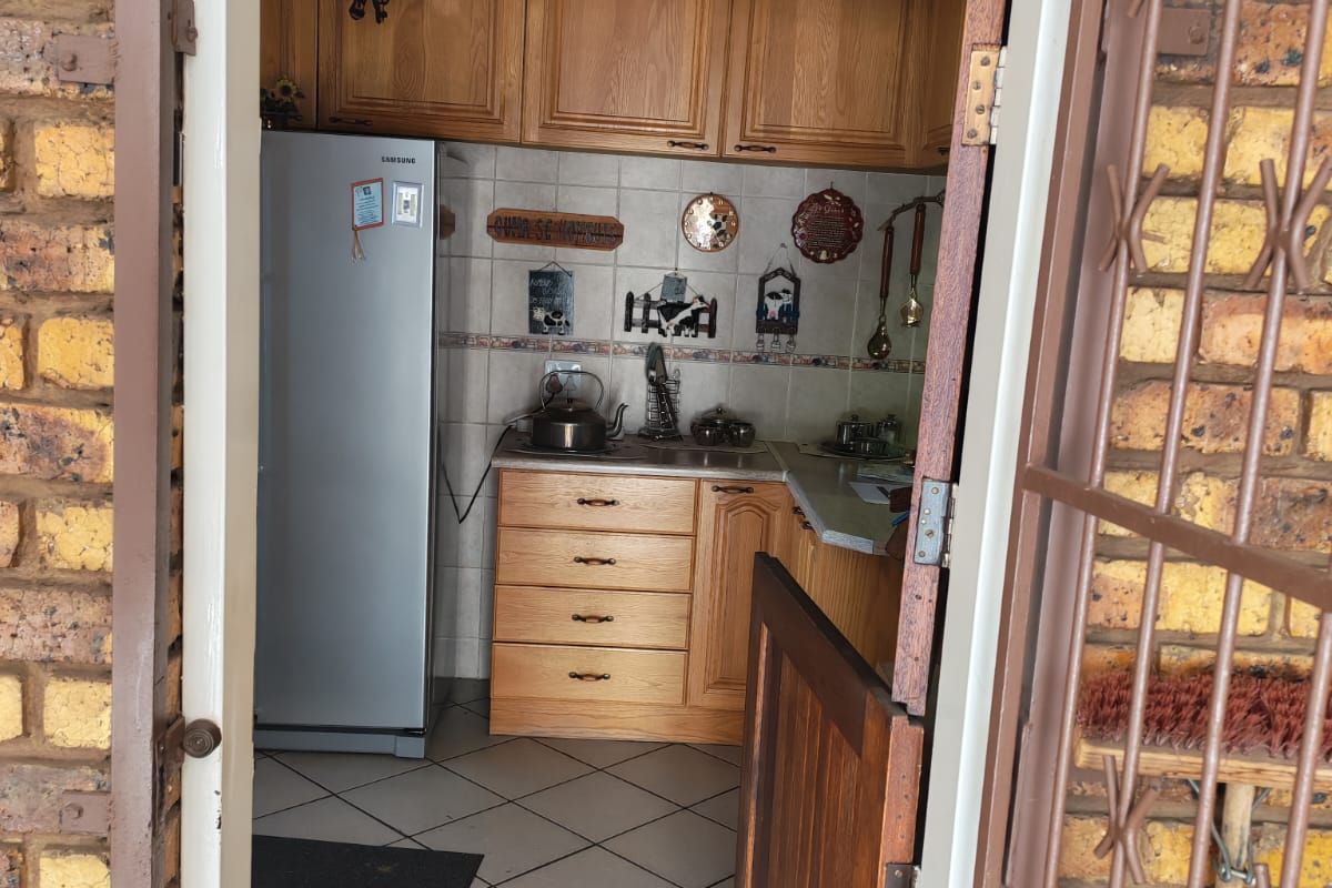 A compact kitchen featuring timber cabinetry, tiled flooring, and a tiled backsplash, viewed through an exterior doorway with security gate.