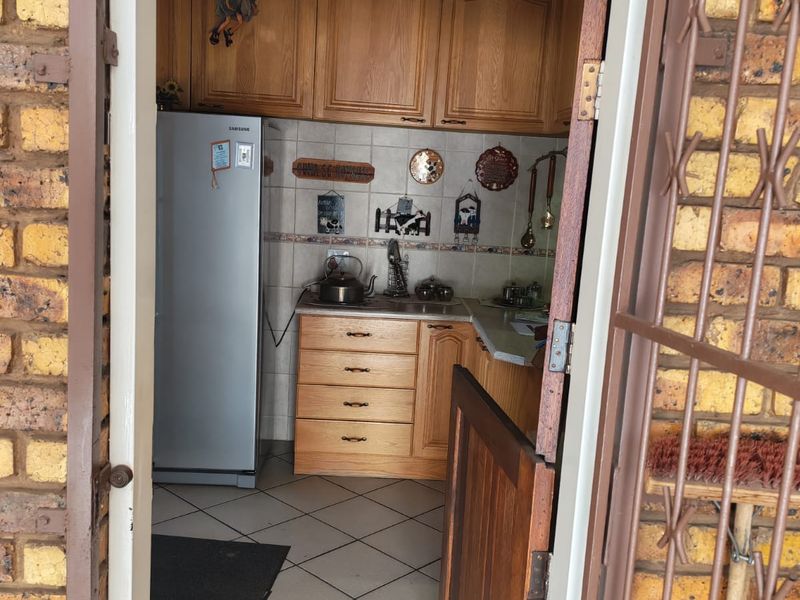 A compact kitchen featuring timber cabinetry, tiled flooring, and a tiled backsplash, viewed through an exterior doorway with security gate.