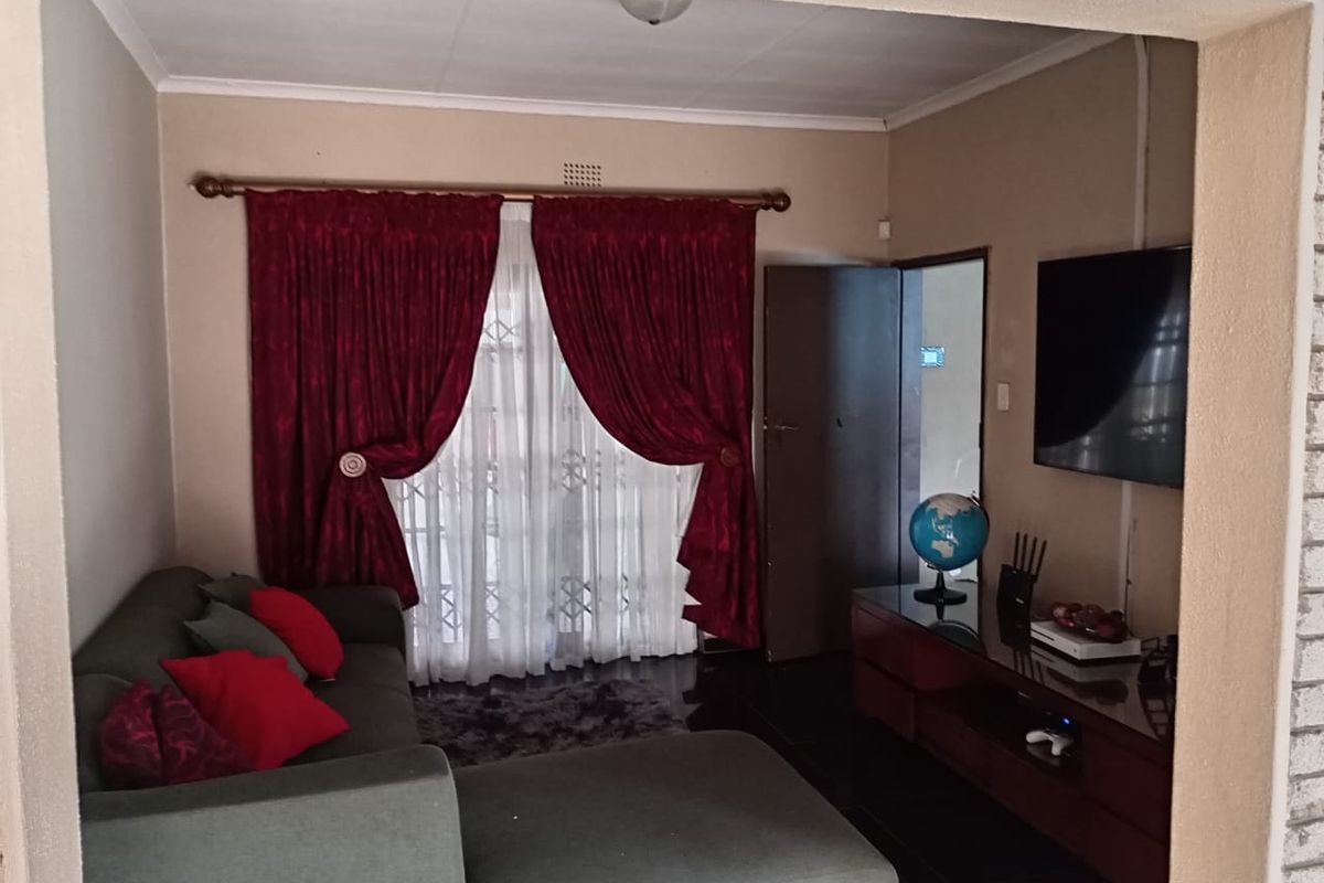 A cozy living room featuring tiled flooring, a large window with decorative curtains, and a wall-mounted television setup.