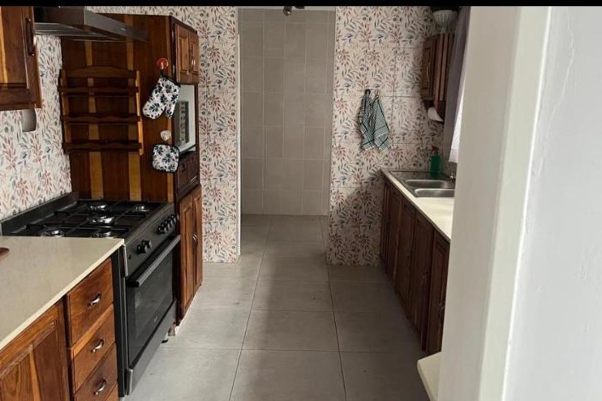 A dated kitchen featuring wooden cabinetry, a gas stove with extractor fan, and tiled flooring, leading into a tiled utility or scullery area.