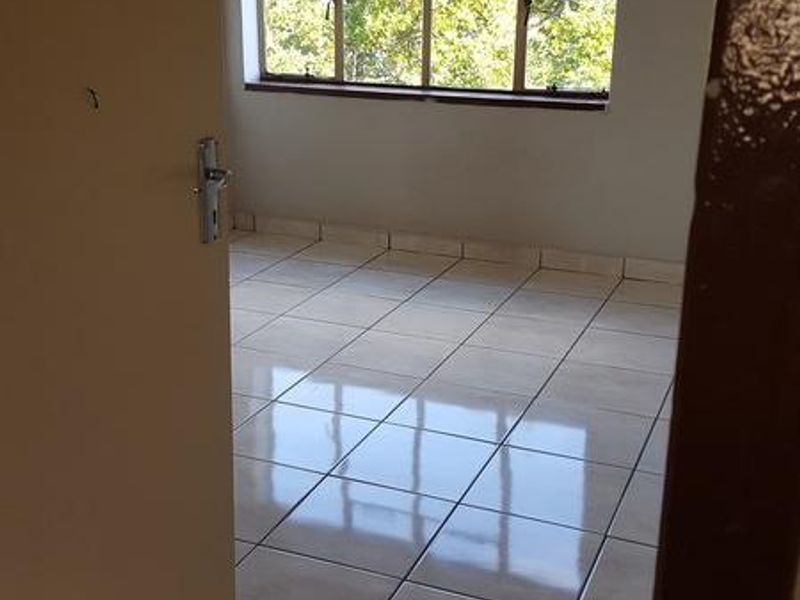 A bright, empty bedroom featuring neutral tiled flooring and a large window providing natural light and a pleasant outdoor view.
