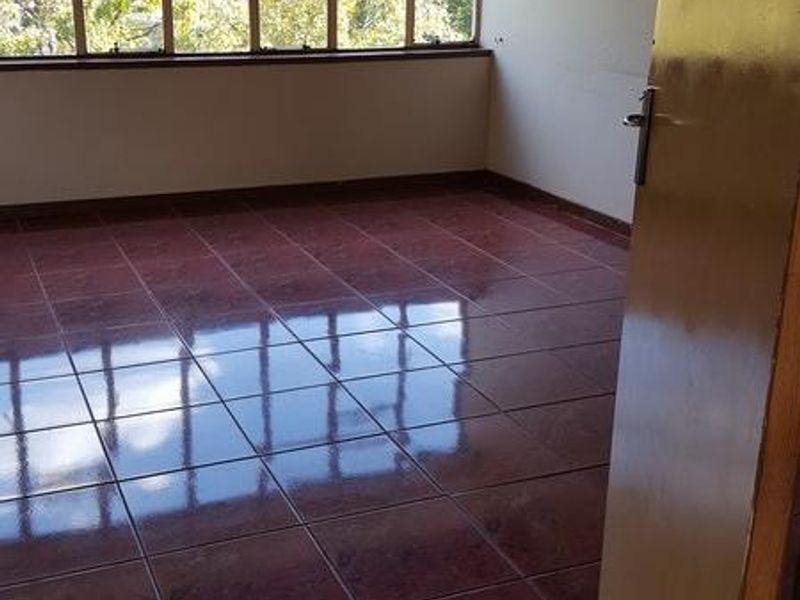 An empty bedroom featuring polished red tiled flooring and a large window providing natural light and a view of the surrounding trees.