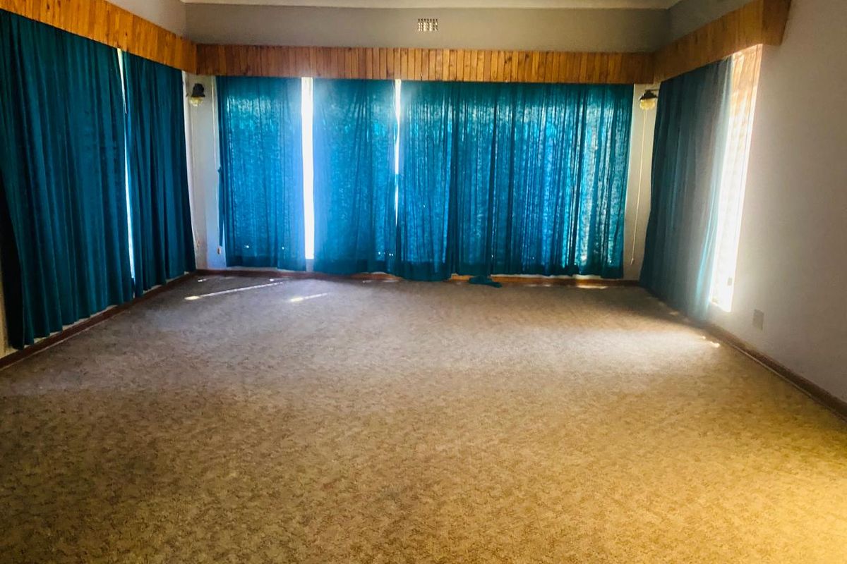 A spacious, empty living room featuring wall-to-wall carpeting, large windows with floor-to-ceiling curtains, and traditional timber pelmet detailing.