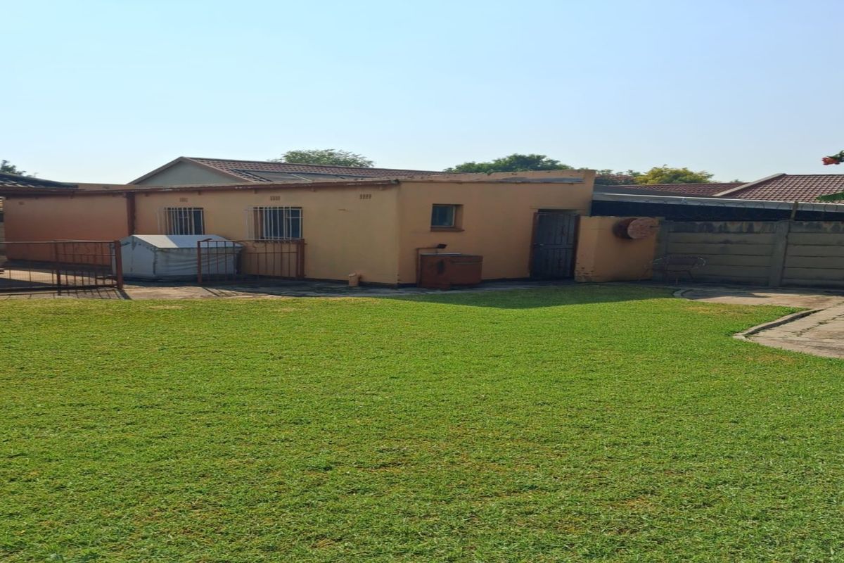 A view of the rear exterior of the house, featuring a large lawn, concrete paving, and a single-story structure with security bars on windows.
