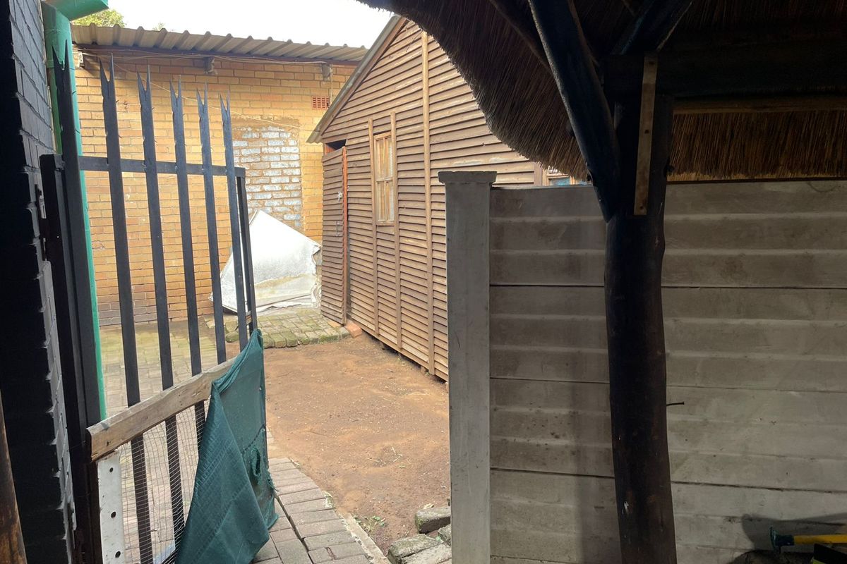 A rear outdoor area featuring a timber structure, a thatched roof section, and paved pathways enclosed by a metal security fence.