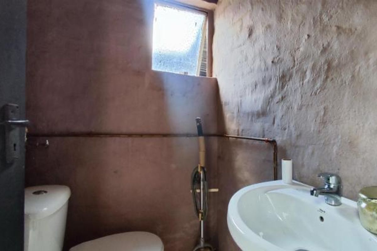A compact bathroom featuring a pedestal basin, toilet, and small window, characterized by rustic, unfinished wall textures and exposed plumbing.