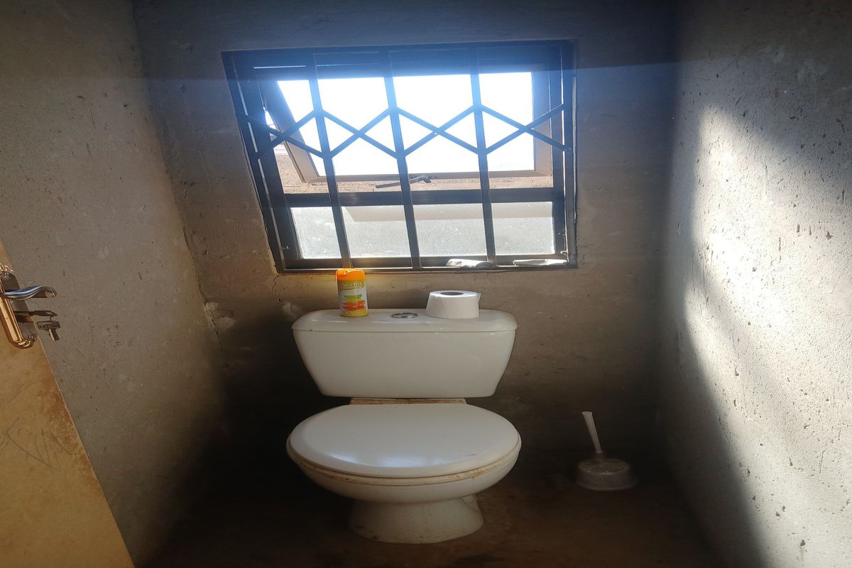A small bathroom featuring a standard toilet and a window with security bars, currently in an unfinished state with exposed concrete walls.