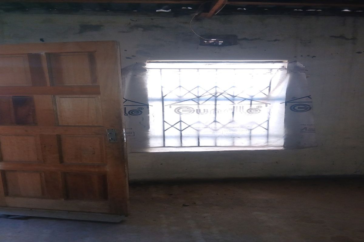 An interior room in poor condition featuring a wooden door, a window with security bars, and exposed electrical wiring on the wall.