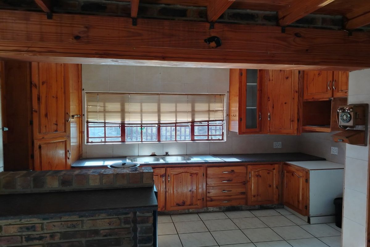 A rustic kitchen featuring timber cabinetry, exposed wooden ceiling beams, tiled countertops, and a double basin sink with a window view.