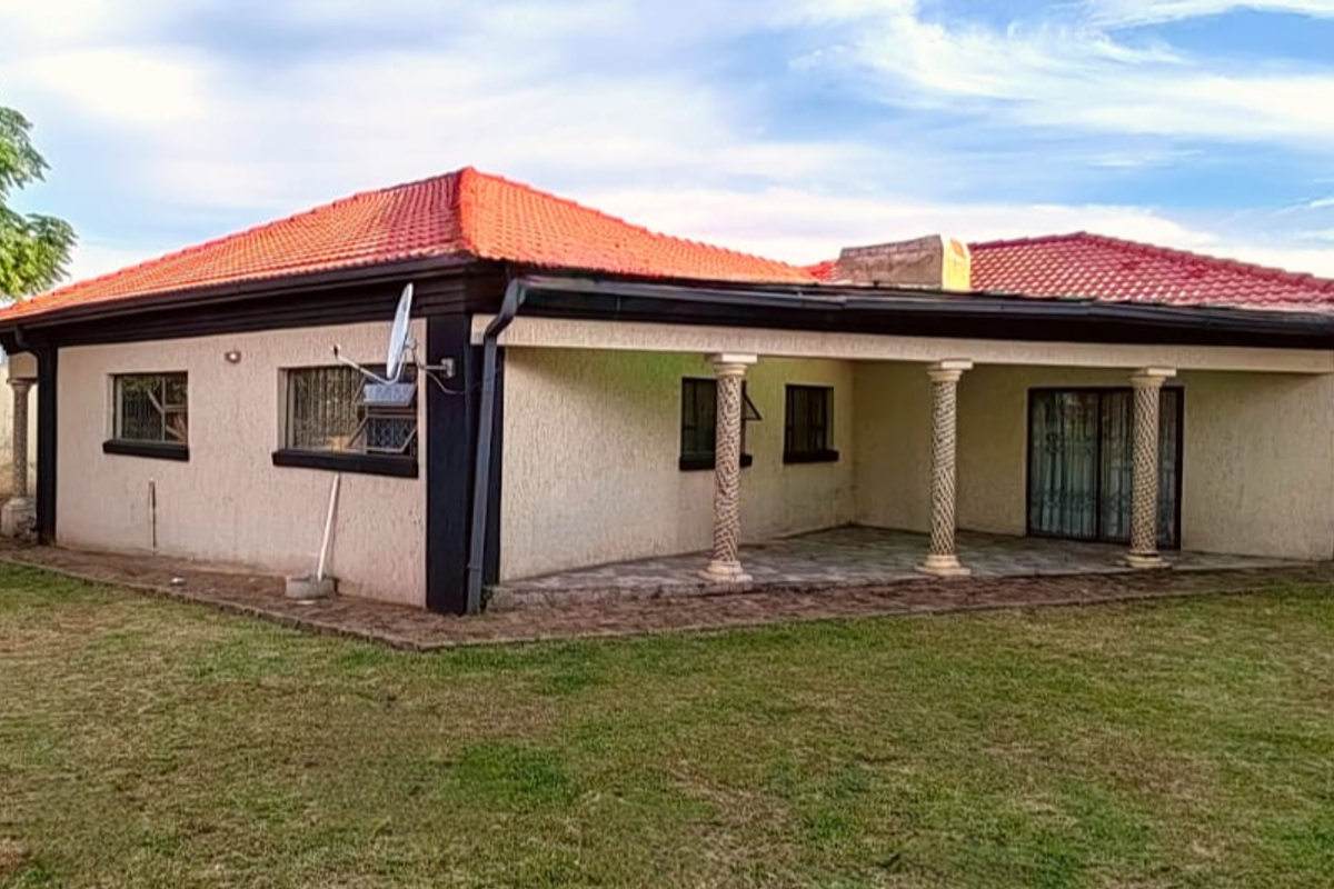 A rear view of a single-story house featuring a covered patio with decorative columns, a tiled roof, and a surrounding lawn area.