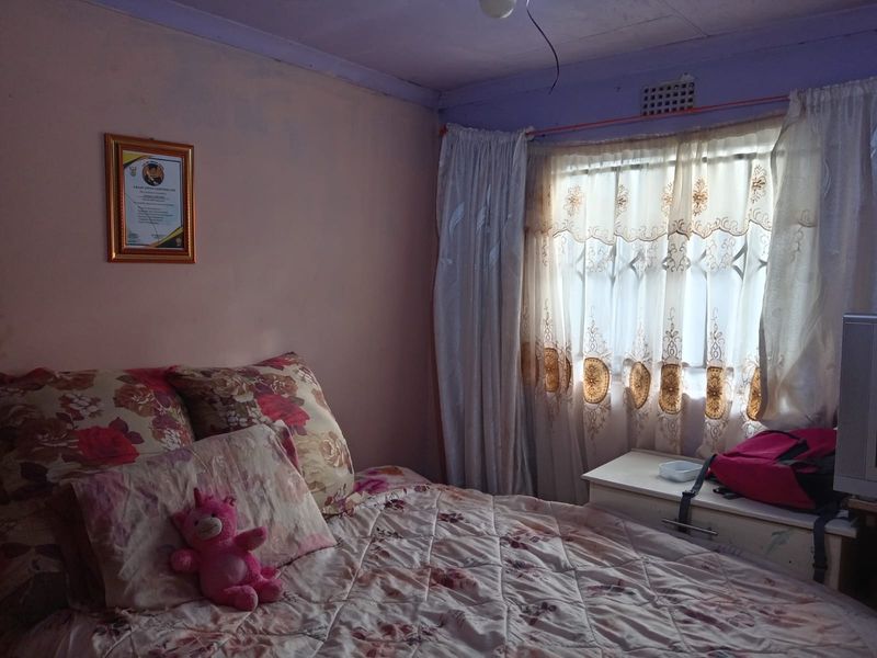 A bedroom featuring a window with security bars and curtains, showing signs of wear on the walls and ceiling.