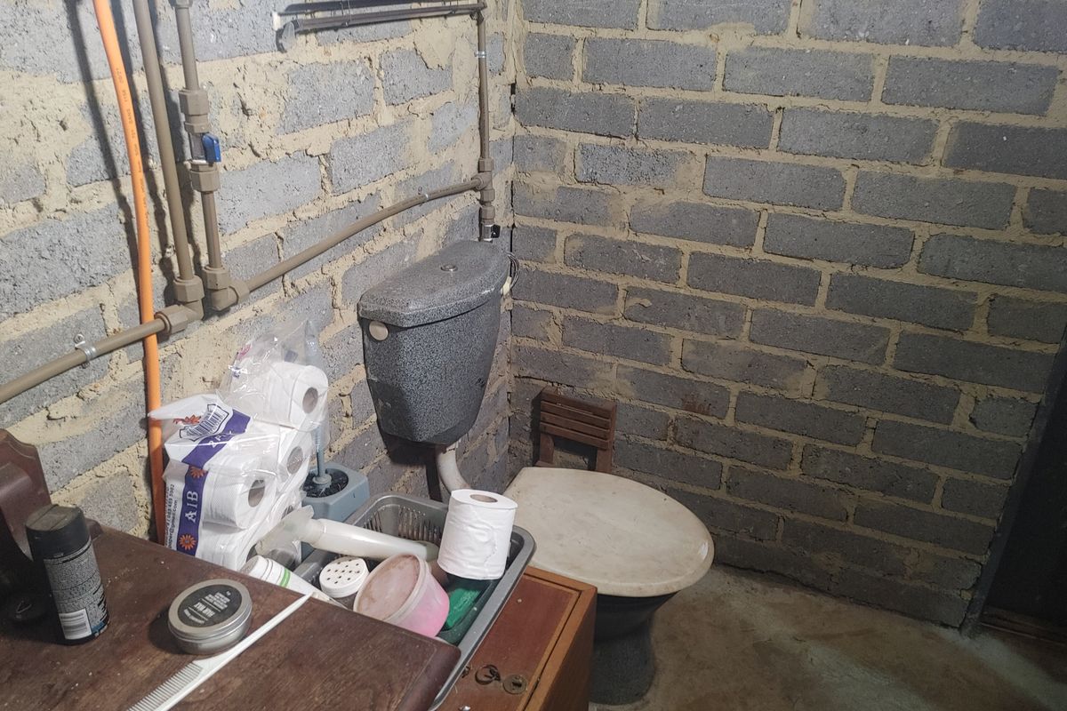 A basic bathroom featuring a toilet with an exposed cistern and visible plumbing against unfinished brick walls and a concrete floor.