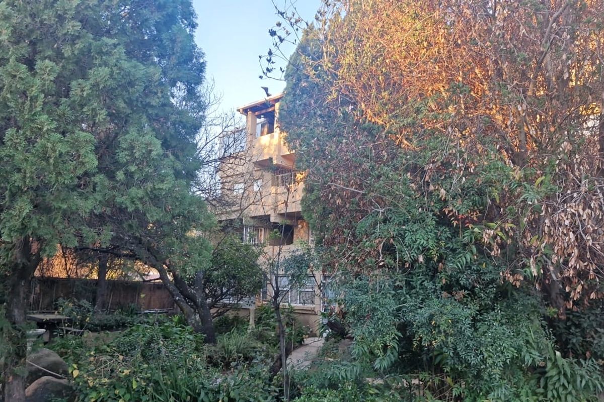 A view of the apartment building exterior, partially obscured by mature trees and garden landscaping.