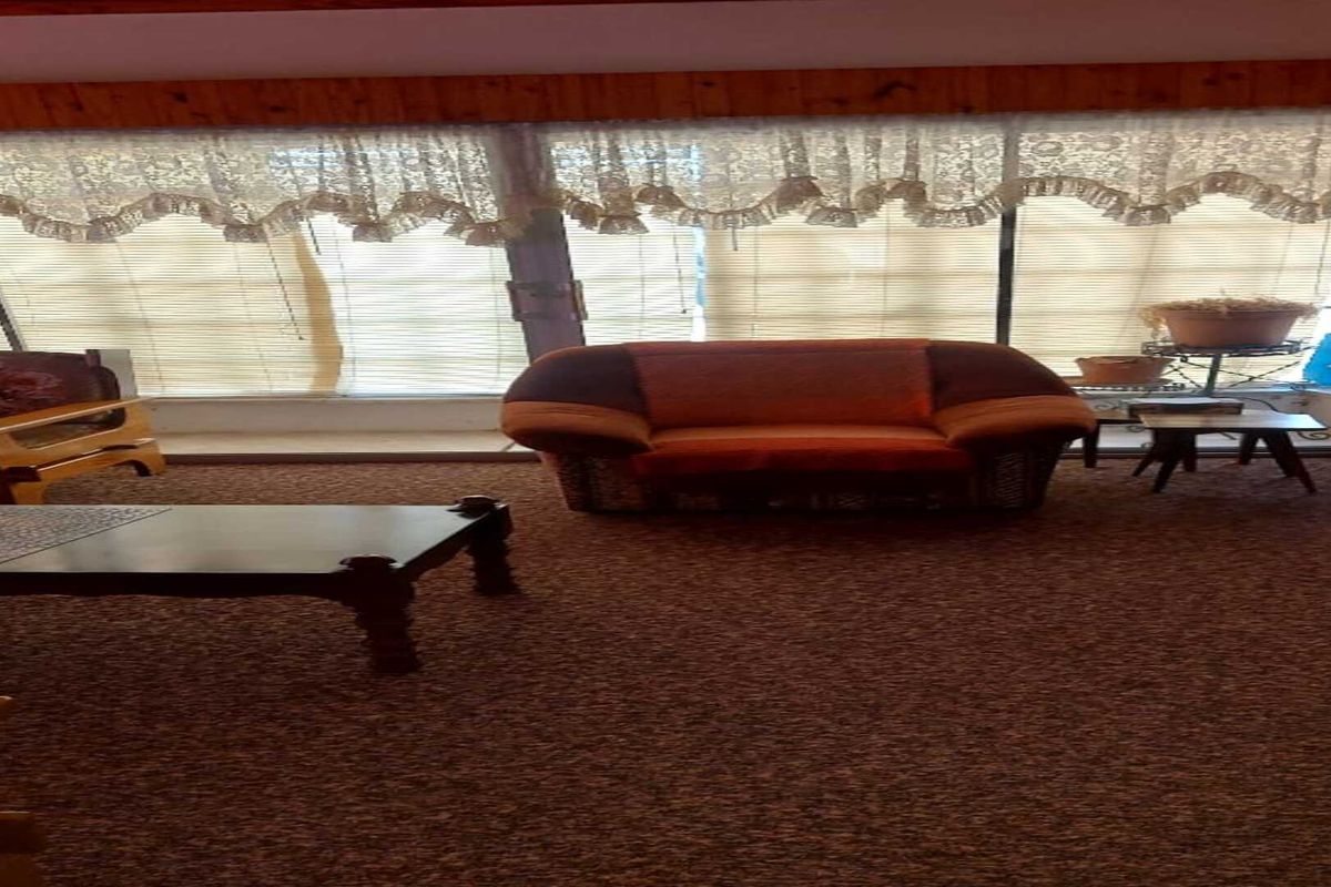A living room featuring wood-paneled ceiling accents and large windows with integrated blinds, offering a dated aesthetic ready for modernization.