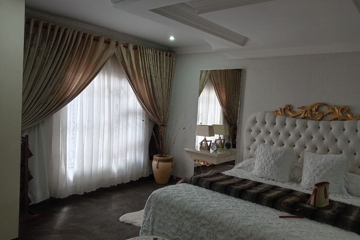 A well-maintained bedroom featuring a coffered ceiling with recessed lighting and large windows dressed with elegant floor-to-ceiling curtains.