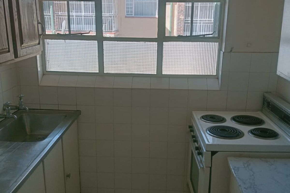 A compact, dated kitchen featuring original cabinetry, a stainless steel sink, and a freestanding electric stove with tiled splashbacks.