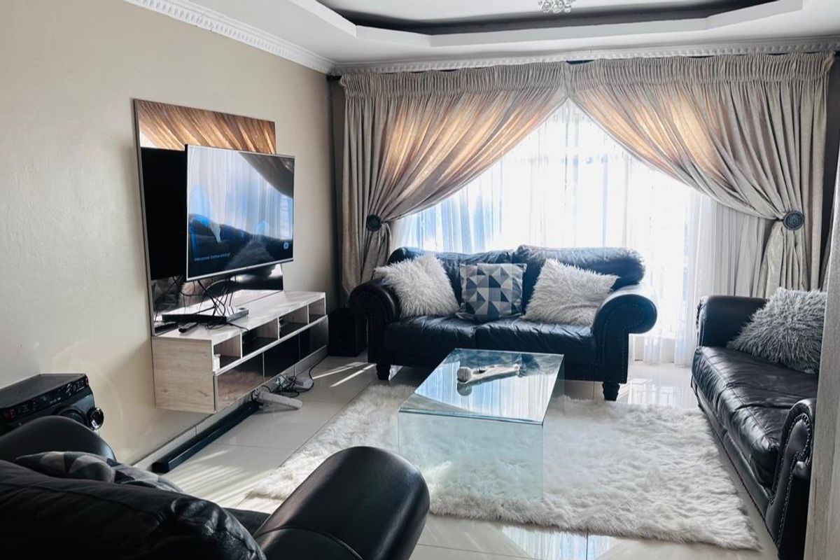 A comfortable living room featuring tiled flooring, a tray ceiling with crown molding, and large windows dressed with floor-to-ceiling curtains.