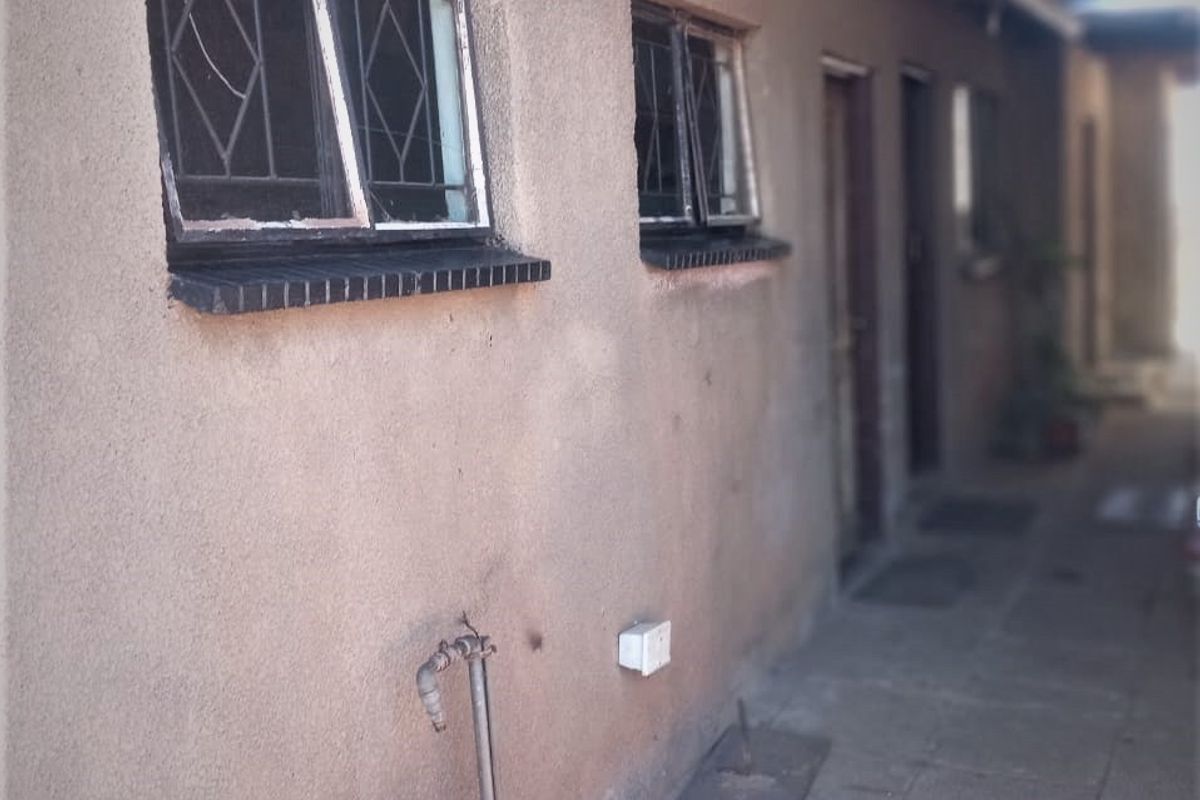Exterior side walkway featuring weathered walls, metal-framed windows with security bars, and basic paved flooring with an external water tap.