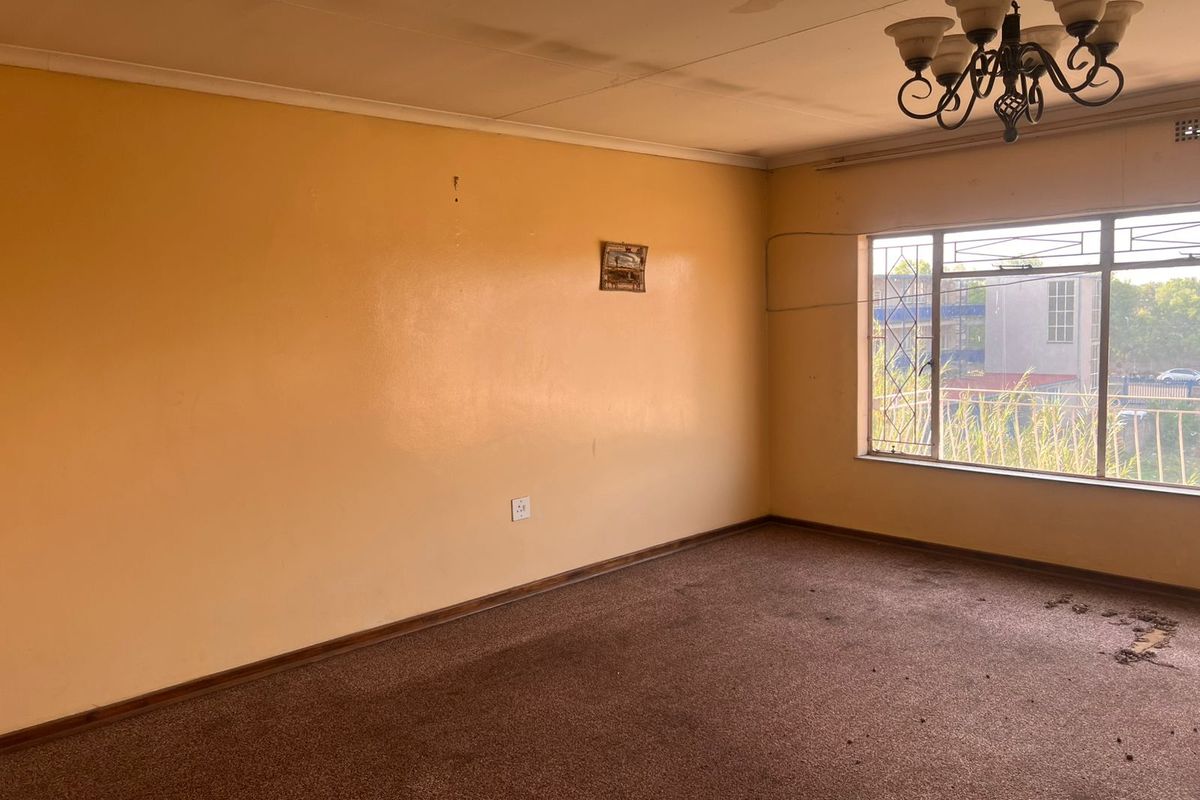 An empty living room featuring brown carpeting, a decorative chandelier, and a large window with security bars, requiring cosmetic updates.