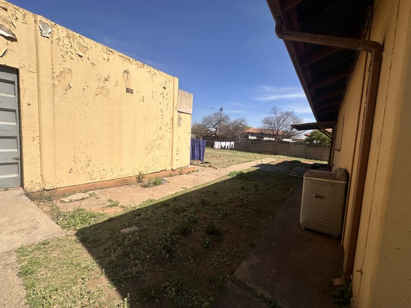 Rear exterior view of the property featuring a paved walkway, lawn area, and boundary wall with visible signs of exterior maintenance required.