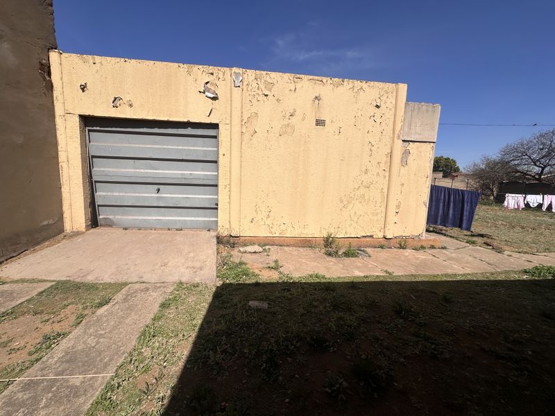 Exterior view of a detached garage featuring a metal roll-up door and concrete driveway, showing significant signs of exterior wall weathering and paint deterioration.