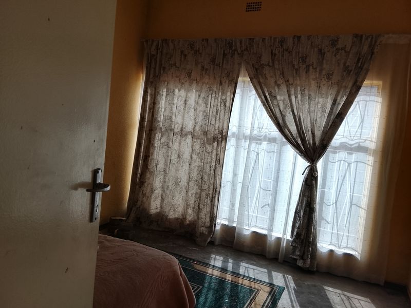 A bedroom featuring natural light through large windows with curtain rails and a security gate, requiring cosmetic updates to the walls and finishes.