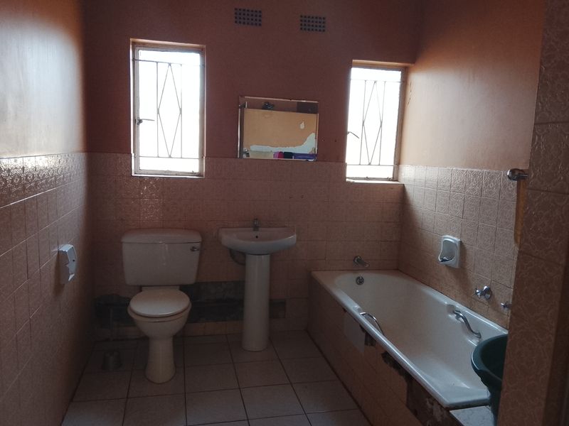 A dated bathroom featuring a pedestal basin, toilet, and bathtub with tiled finishes, requiring cosmetic updates and repairs to wall and bathtub paneling.
