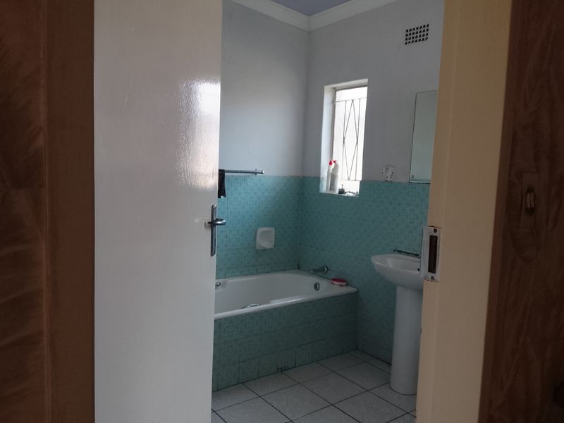 A functional bathroom featuring a bathtub, pedestal basin, and dated blue wall tiling, viewed through an open doorway.